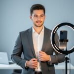 Person in a blazer facing a smartphone on a ring-light tripod in a home office, preparing a professional headshot with soft daylight and a blurred laptop and neutral background.